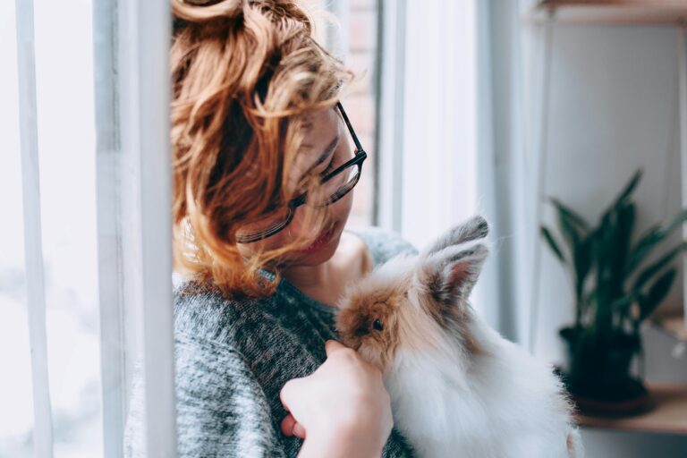 Young woman with glasses cuddling a fluffy rabbit indoors near a window.